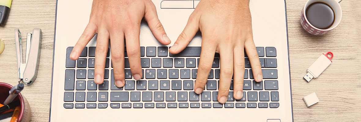 A person’s hands typing on a laptop keyboard, with a stapler, pens, USB drive, and a cup of coffee on the wooden desk.