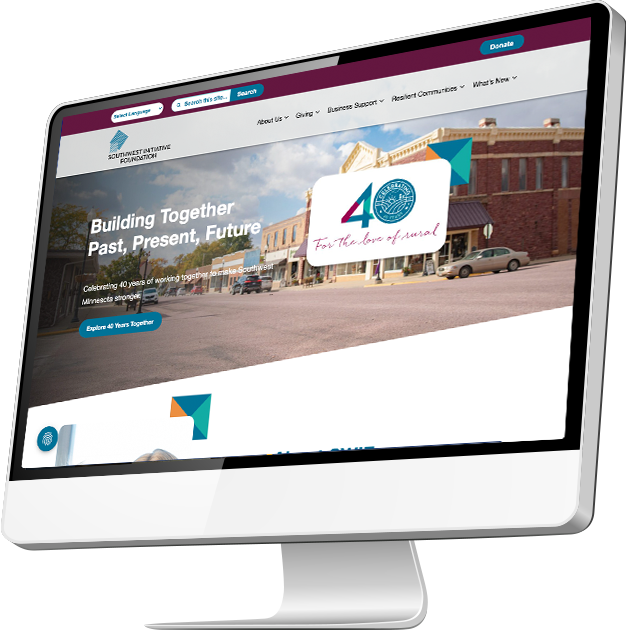 A computer monitor displaying a website with the text Building Together Past, Present, Future and a logo celebrating a 40th anniversary, set against a background of brick buildings on a sunny street.