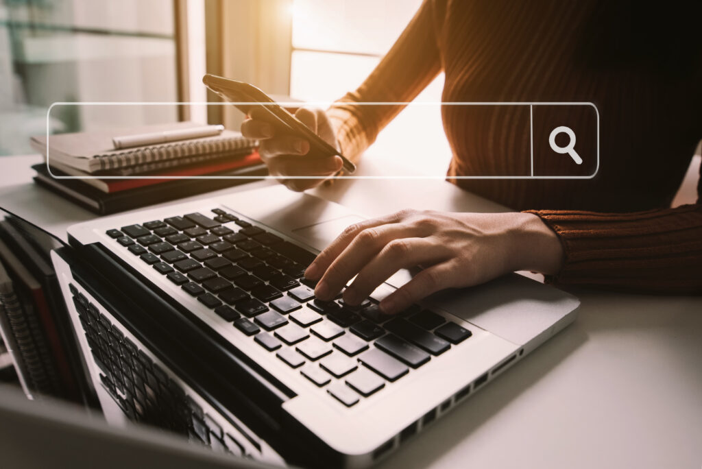 A person is using a smartphone and typing on a laptop keyboard, with a transparent search bar overlay featuring the query “what is a landing page” and a search icon, in a bright office setting.