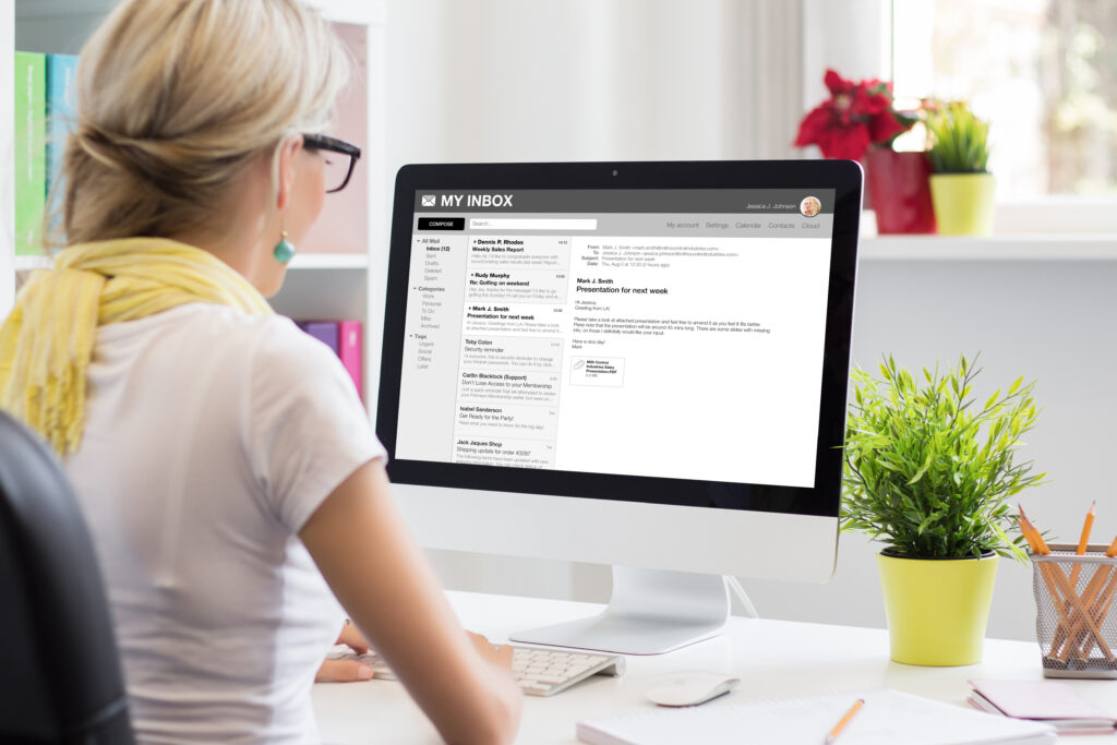 A woman with blonde hair and glasses sits at a desk, looking at mass mailing emails on a large computer monitor. The workspace includes a green plant, notebooks, and shelves with colorful folders in the background.