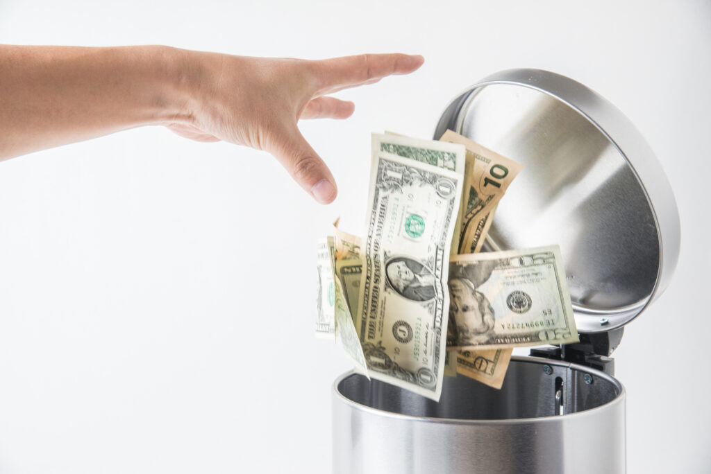 A hand drops several US dollar bills into a silver trash can against a plain white background, symbolizing wasted resources that could have been invested in foundational marketing.