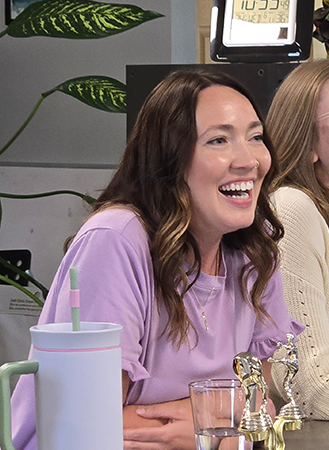 A woman with wavy brown hair, wearing a lavender shirt, laughs while seated at a table adorned with trophies, a tumbler with a straw, and a glass of water—each item featuring playful title tags. Another person sits beside her. Green plants are in the background.