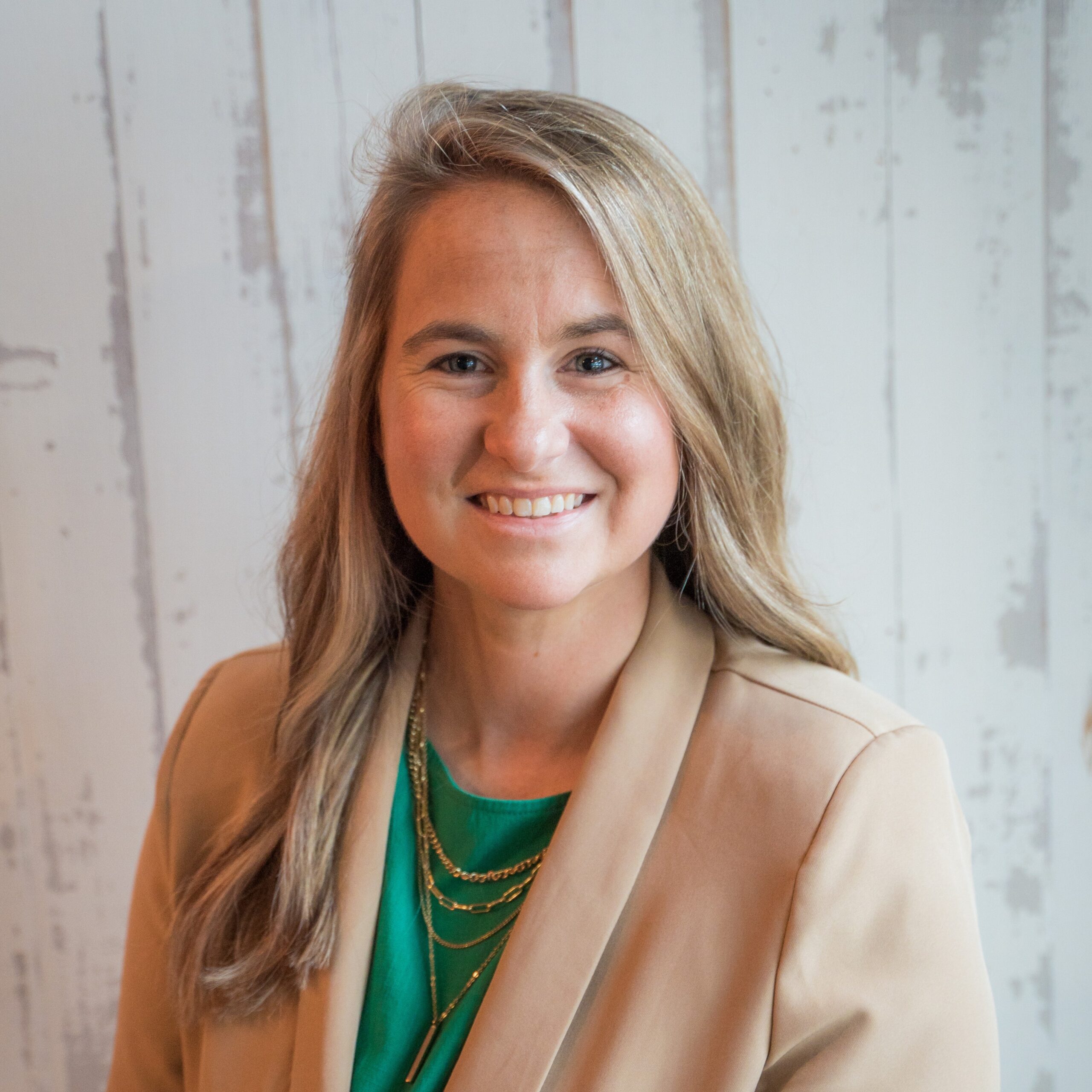 A woman with long, light brown hair smiles at the camera. She is wearing a tan blazer over a green top and layered gold necklaces, standing in front of a white wooden wall with a weathered look.