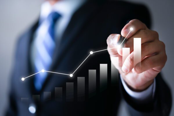 A person in a suit draws a rising line graph with a marker, illustrating growth or success—an ideal image for learning how to grow a referral-based business over a blurred bar chart background.