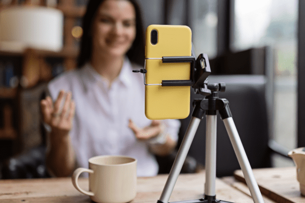 A woman sits at a table with a coffee mug, speaking and gesturing towards a smartphone mounted on a tripod, possibly recording a video or video calling. The background is blurred with bookshelves and a lamp.