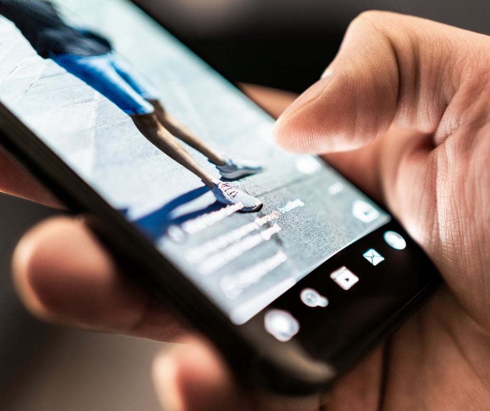 Close-up of a person using their right thumb to scroll through a social media feed on a smartphone, with a short-form video for business featuring someone in shorts and sneakers displayed on the screen.