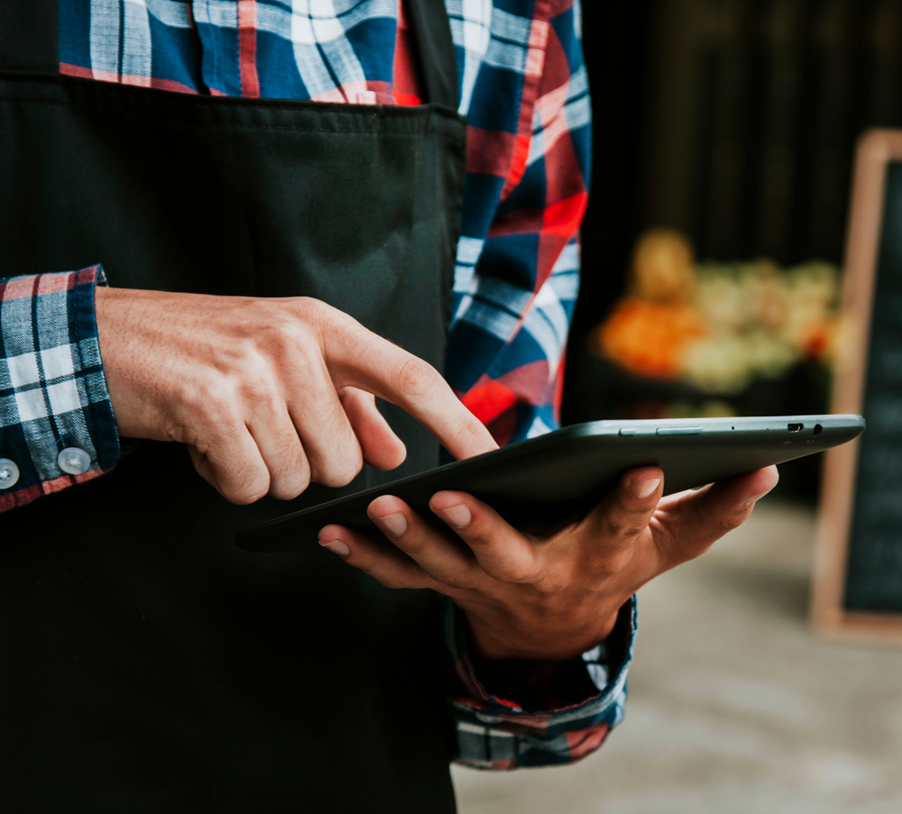 A person wearing a plaid shirt and black apron uses a tablet, tapping the screen with one finger. The background is blurred, suggesting an indoor or shop setting.