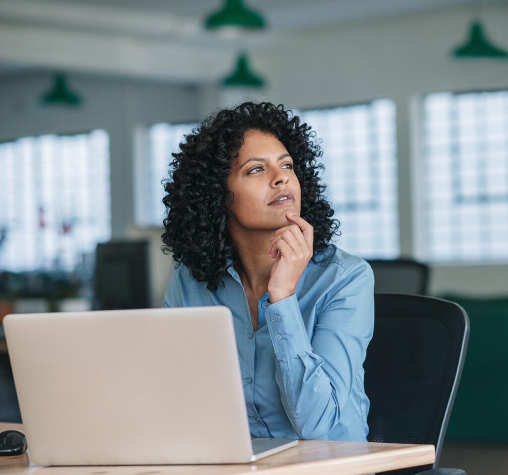A person with curly hair wearing a blue shirt sits at a desk with a laptop, pondering the importance of a website for AI, their hand resting on their chin as they gaze thoughtfully into the distance in a bright, modern office.