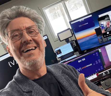 A smiling man with glasses and gray hair gives a thumbs up in front of multiple computer monitors displaying virtual meeting and digital content in a bright home office.