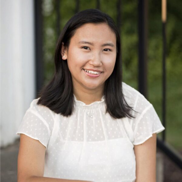 A young woman with straight black hair and a white, short-sleeved, semi-sheer polka dot blouse sits outdoors, smiling at the camera—perhaps taking a break from her marketing team—with greenery and a black railing in the background.