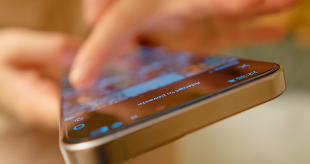 A close-up of a persons hand using a smartphone, with fingers interacting with the touchscreen. The phone display is slightly blurred, showing app icons and notifications.