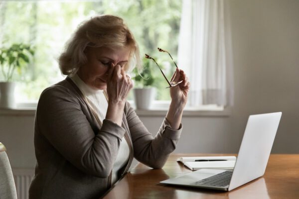 An older woman sits at a desk with a laptop and notebook, holding her glasses in one hand and rubbing her eyes with the other, looking tired or stressed. Light filters in through a window behind her.