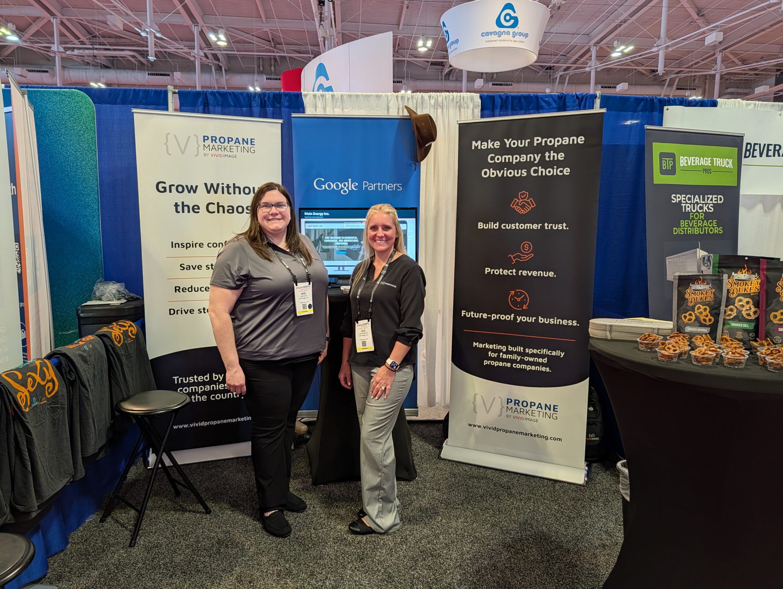 Two women stand smiling at a trade show booth for Propane Marketing Group. The booth displays banners about marketing services, a screen with Google Partners, and containers of pretzels on a table.
