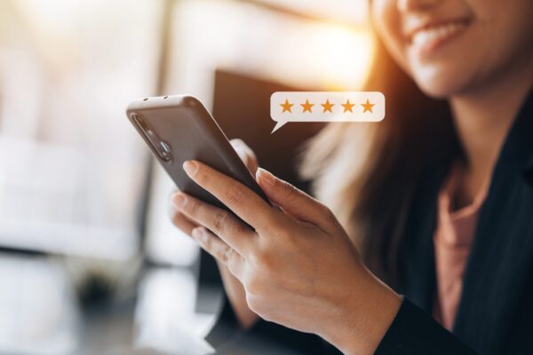 A smiling woman holds a smartphone, with a digital illustration of a speech bubble showing five gold stars, suggesting she is viewing or leaving positive Google reviews for local businesses.