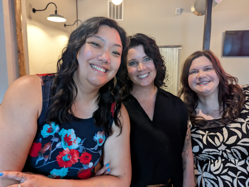 Three women smile at the camera indoors. The woman on the left wears a navy dress with red floral patterns, the center woman wears a black top, and the woman on the right wears a black and white patterned dress. Warm lighting and wall decor are visible.