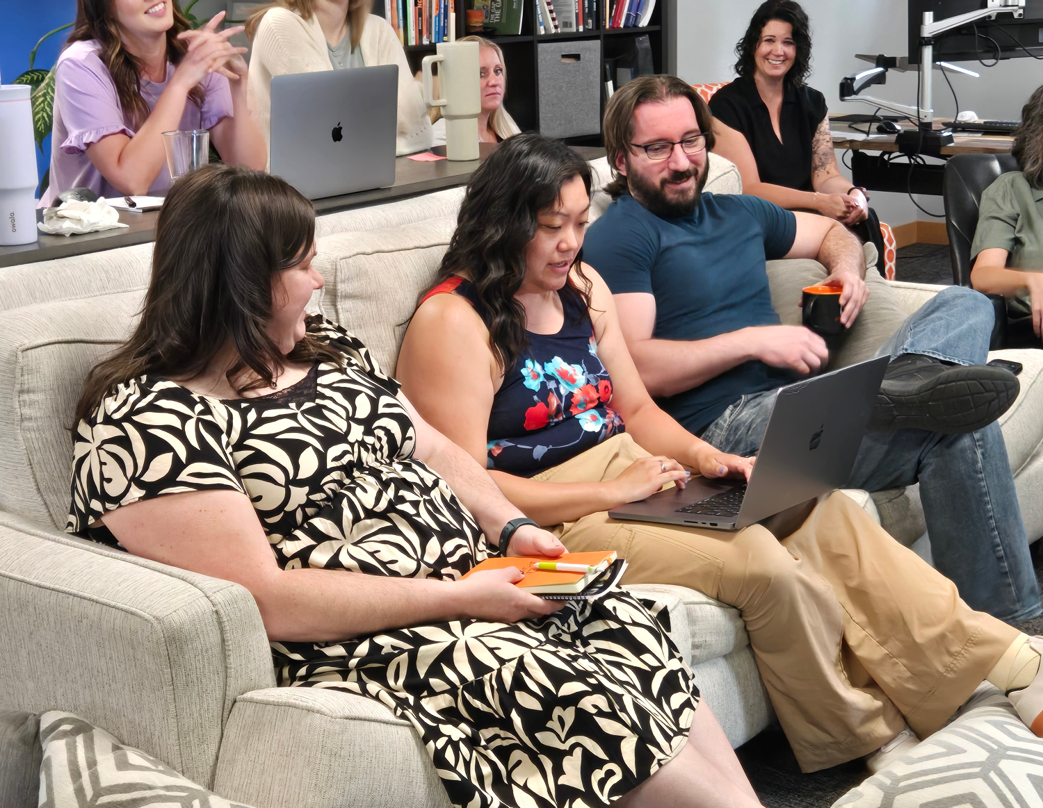 A group of people sit on a couch and chairs in a virtual marketing company office, some holding mugs and laptops, appearing engaged in conversation or a meeting. Bookshelves and computers are visible in the background.