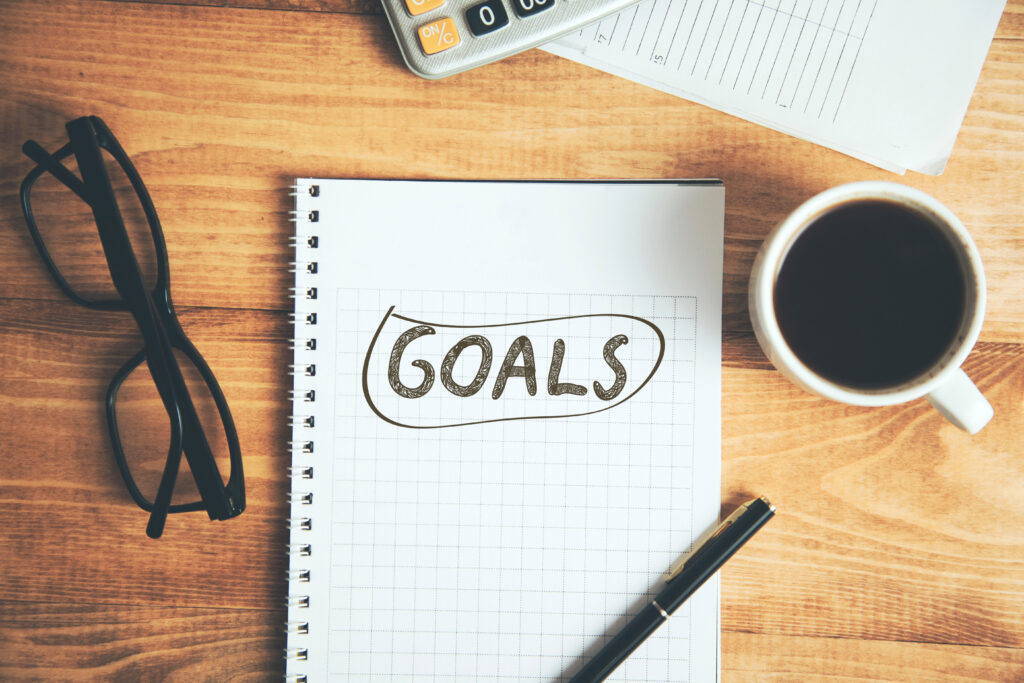 A notebook with “GOALS” written on graph paper, placed on a wooden desk beside a pair of glasses, a black pen, a cup of coffee, a calculator, and some documents.