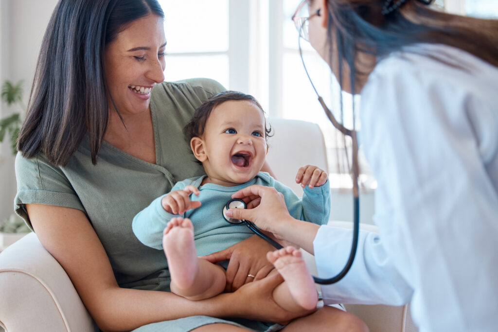 A smiling mother holds her happy baby while a doctor listens to the babys chest with a stethoscope during a check-up in a brightly lit room.