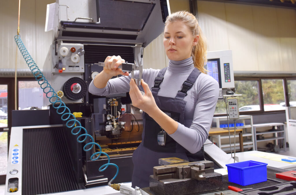 A woman in overalls measures a metal part with a tool in a modern industrial workshop, surrounded by machinery and equipment.