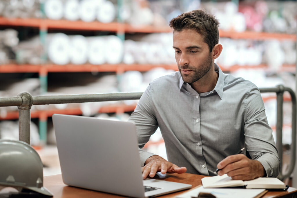 A man in a gray shirt works on a laptop at a desk in a warehouse, taking notes in a notebook. Shelves with large rolls of material are visible in the background. A gray hard hat sits on the desk nearby.