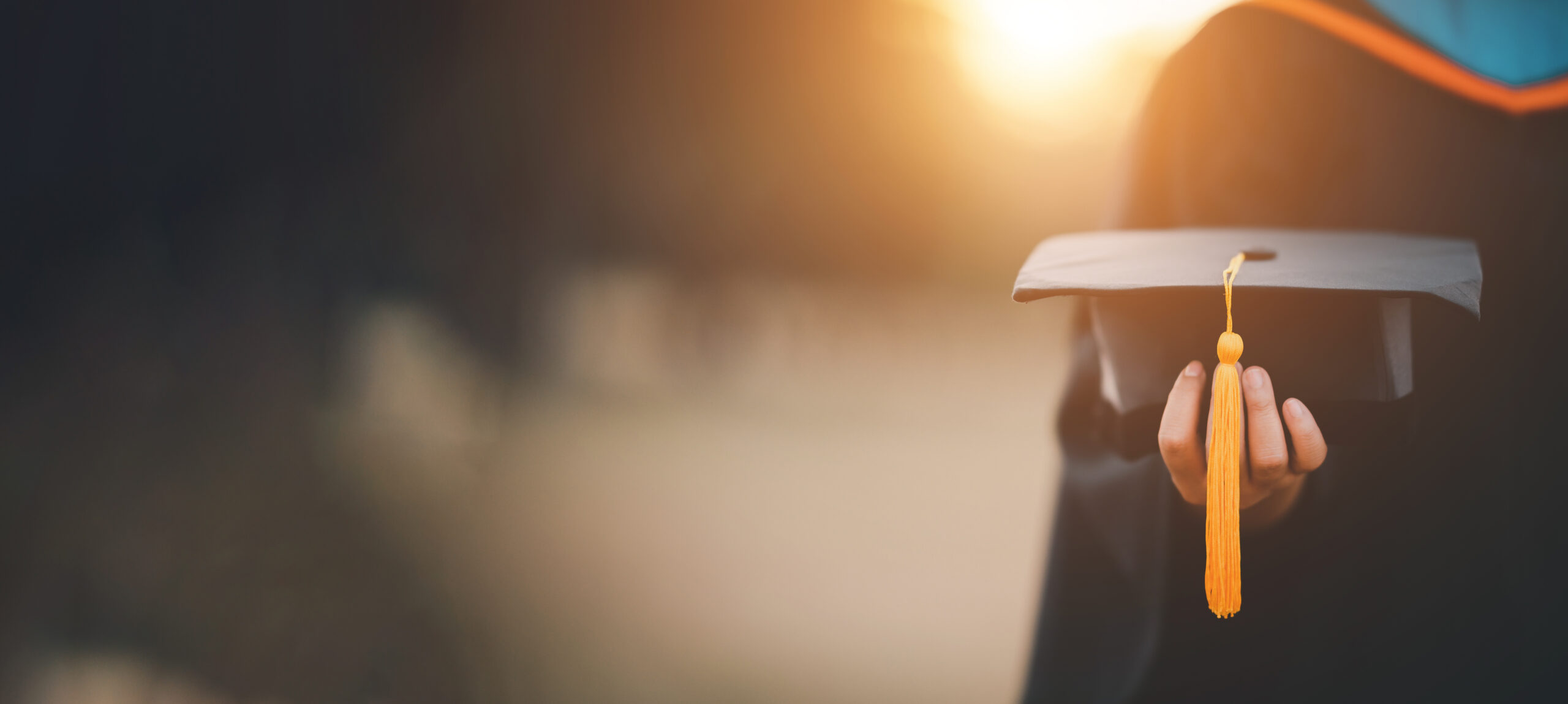 A person in a graduation gown holds a graduation cap with a yellow tassel, with sunlight shining in the background.