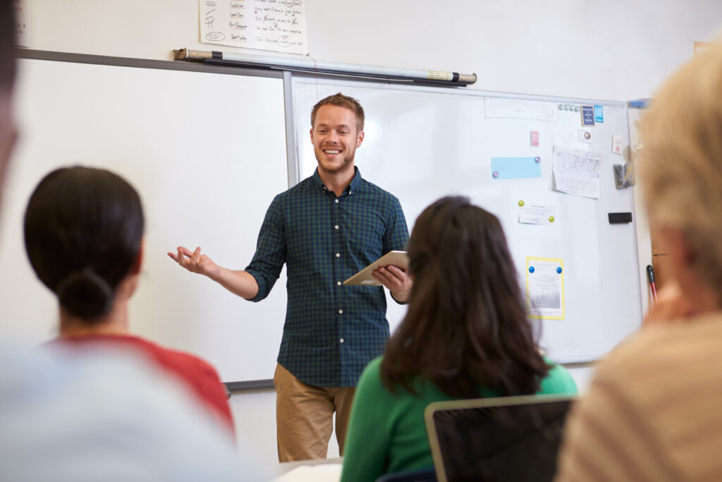 A man stands in front of a whiteboard holding a tablet, smiling and speaking to a group of people seated and listening in a classroom setting.