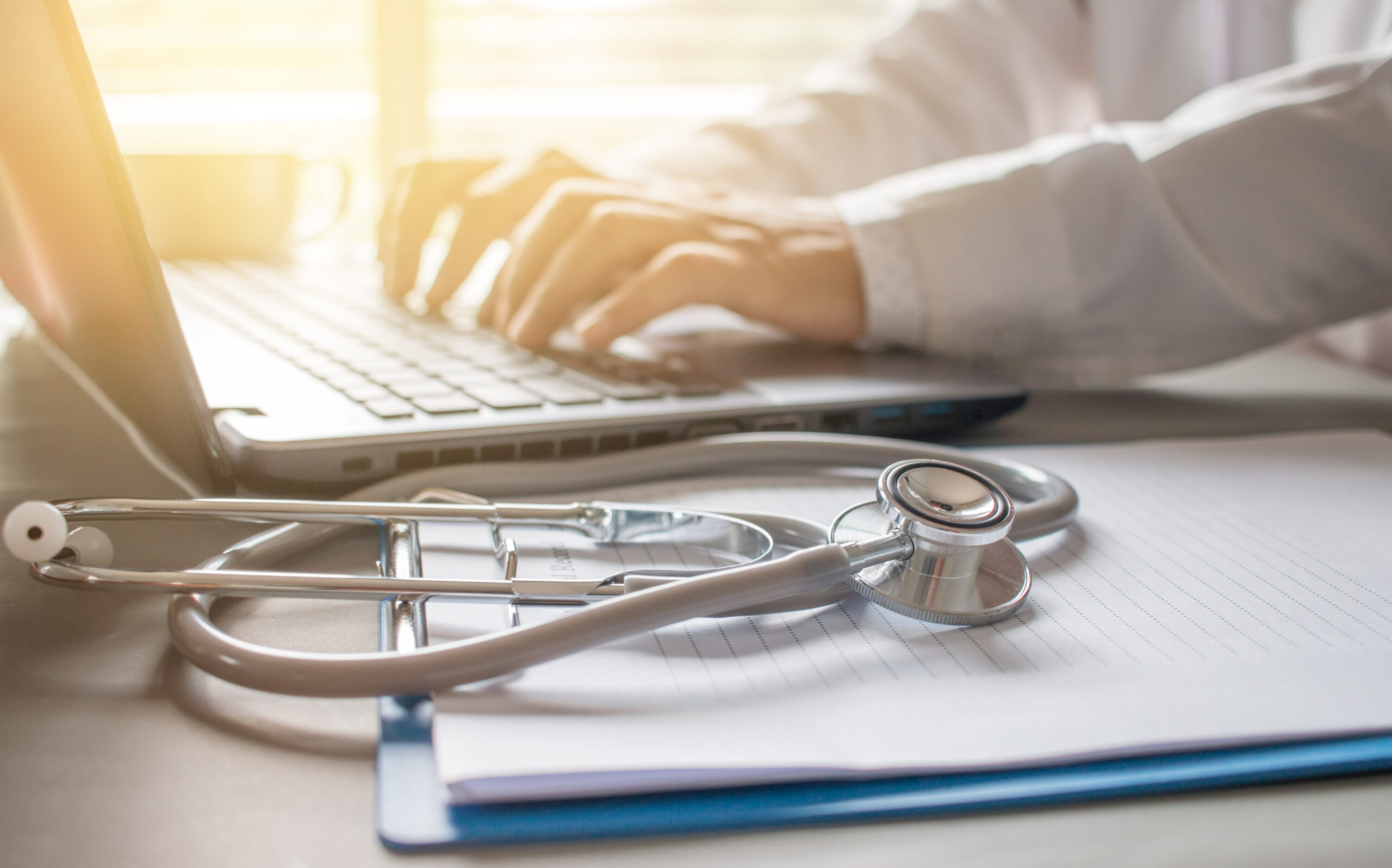 A person in a white coat types on a laptop beside a stethoscope and a clipboard with paper, suggesting a medical or healthcare setting.