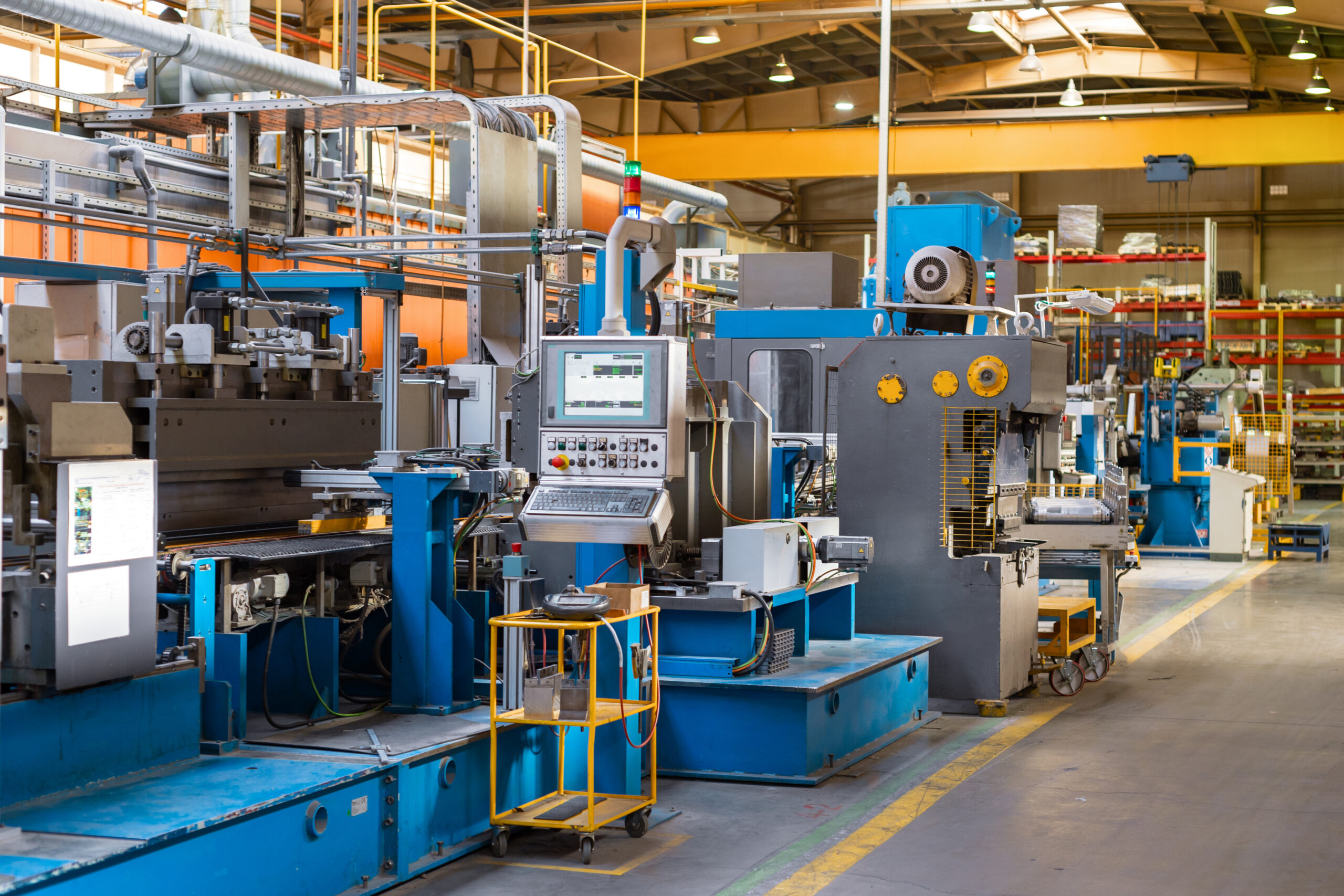 A brightly lit factory floor with various large blue industrial machines, computer monitors, and control panels, surrounded by pipes and organized metal shelving.