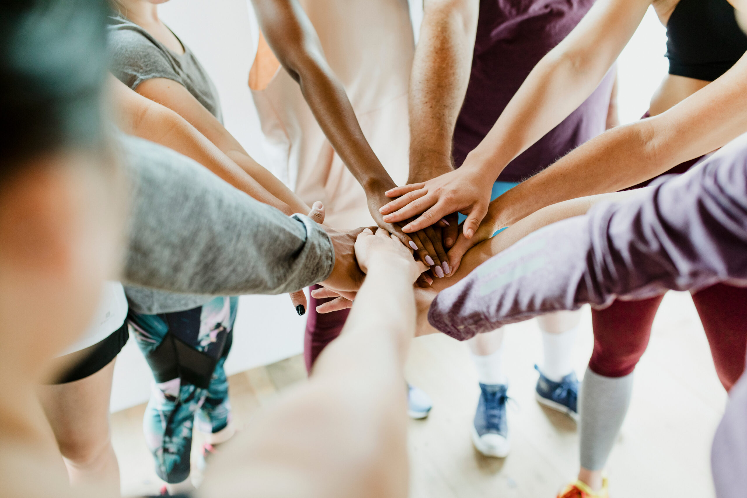 A group of people stand in a circle, reaching their hands into the center, stacking them together in a gesture of teamwork and unity.