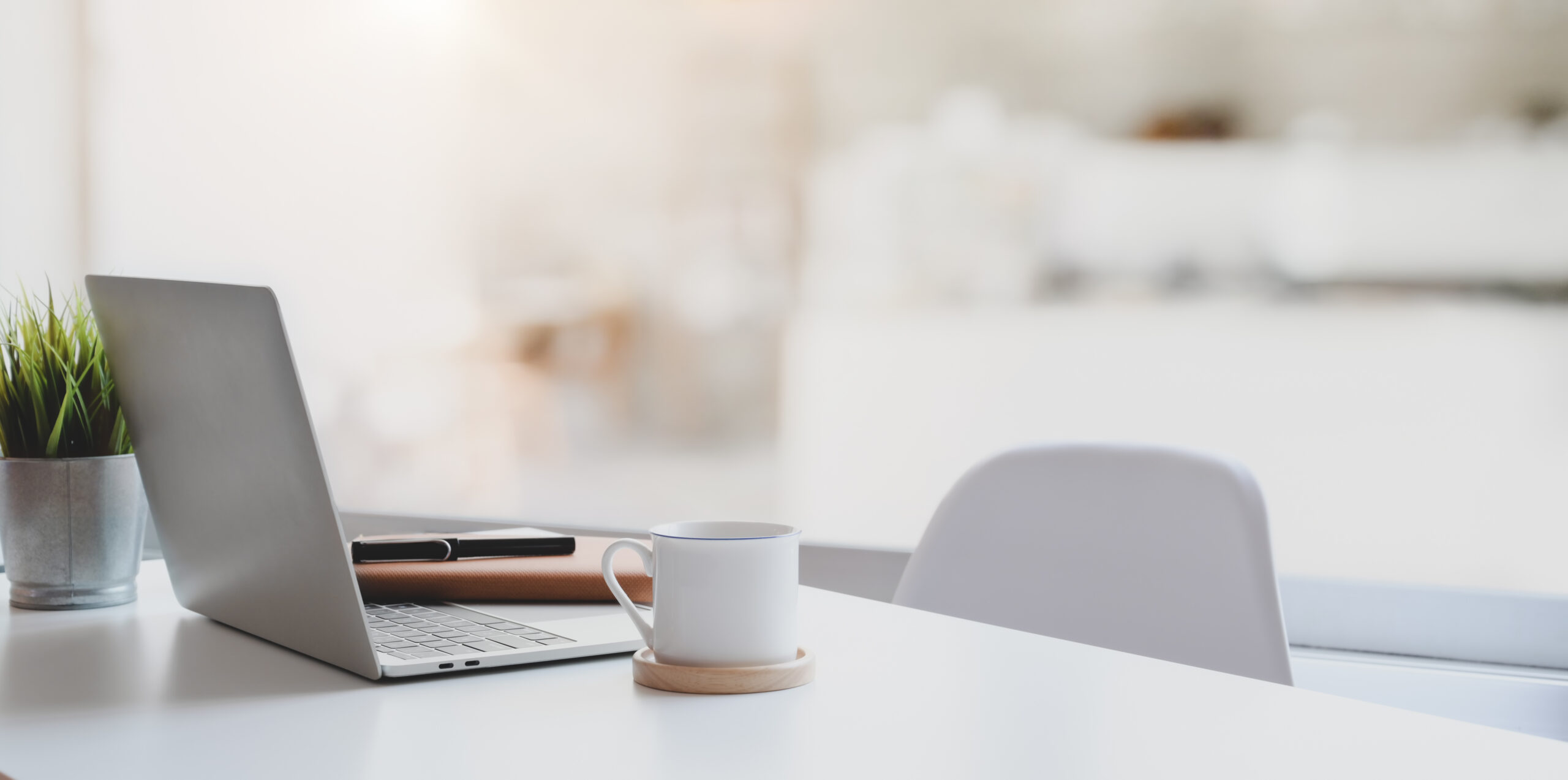 A silver laptop, a notebook, a pen, a white mug on a coaster, and a small green plant sit on a white desk with a white chair in a bright, blurred background.