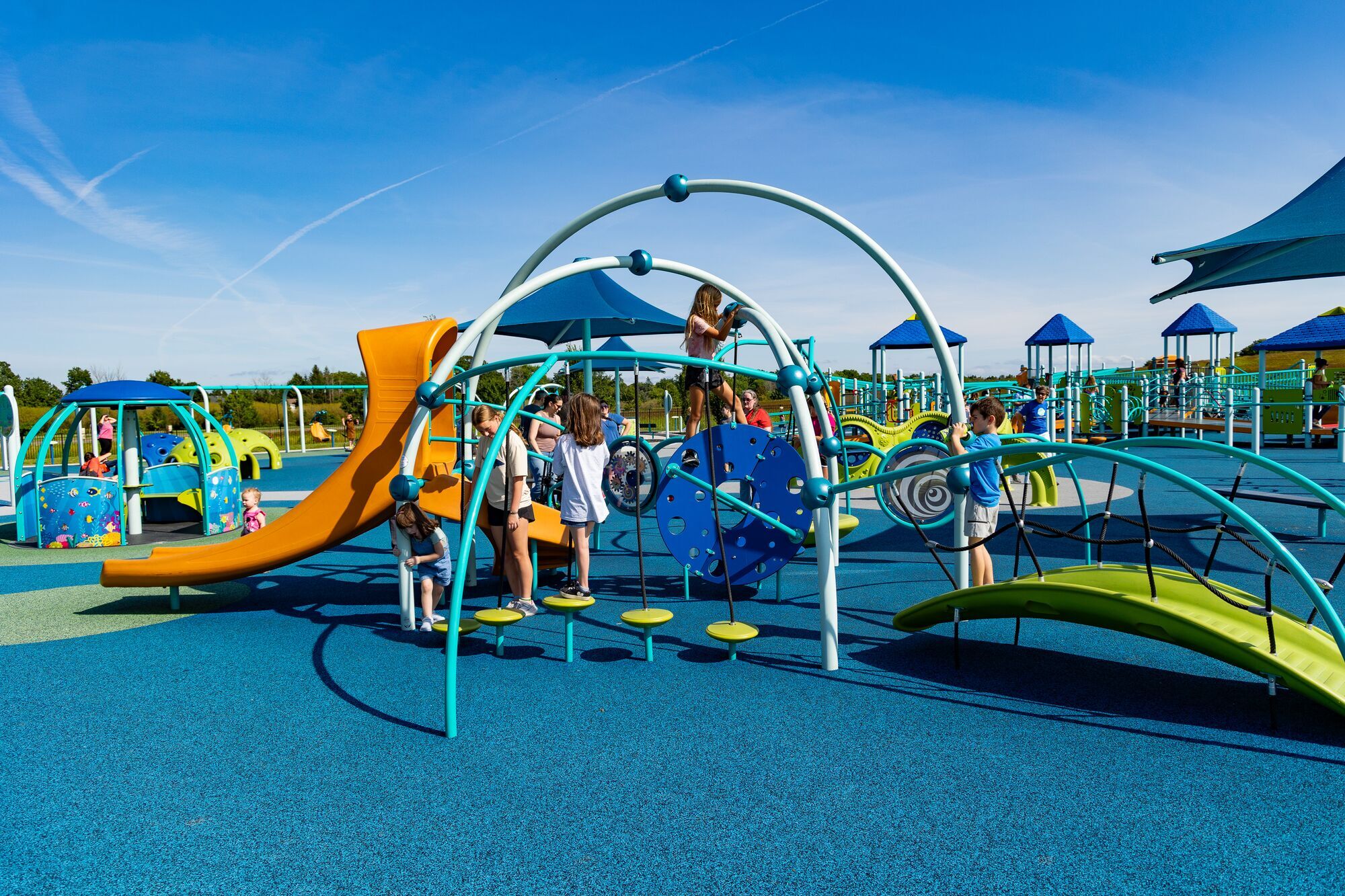 Children play on colorful playground equipment including slides, climbing structures, and tunnels. The ground is covered in blue rubber flooring, and the sky is clear and sunny. Some shade structures are in the background.