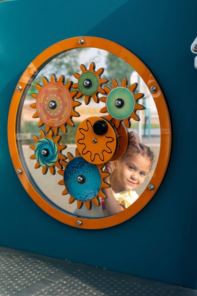 A young child smiles through a transparent panel with colorful, spinning gears mounted on a playground wall. The gears are orange, green, and blue, and the child appears curious and playful behind the display.