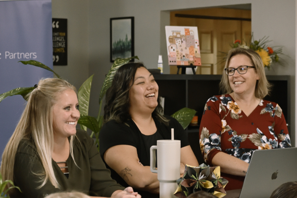 Three women sit at a table, smiling and laughing together. One has a laptop in front of her, and a large drink cup is on the table. The background includes plants, artwork, and a shelf with books.
