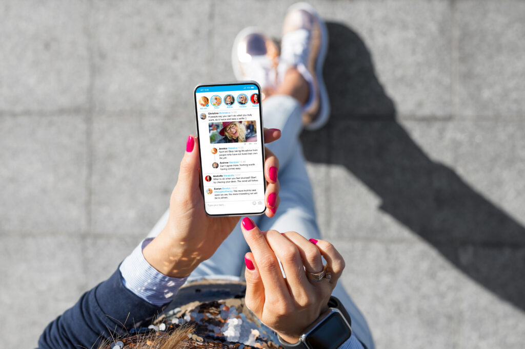 Person holding a smartphone outdoors, scrolling through a social media feed with stories at the top. The person has pink nail polish and is wearing jeans, a blue top, and sneakers.