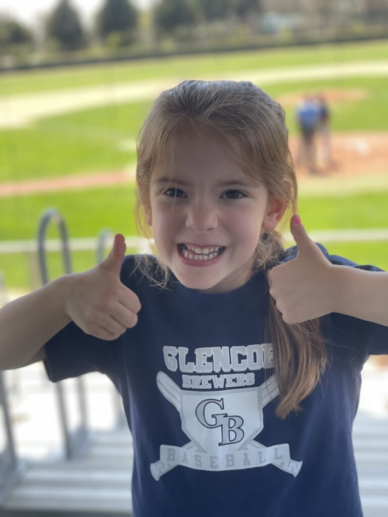 Smiling young girl gives two thumbs up, wearing a Glencoe Braves Baseball T-shirt at an outdoor baseball field, with players visible in the blurry background.