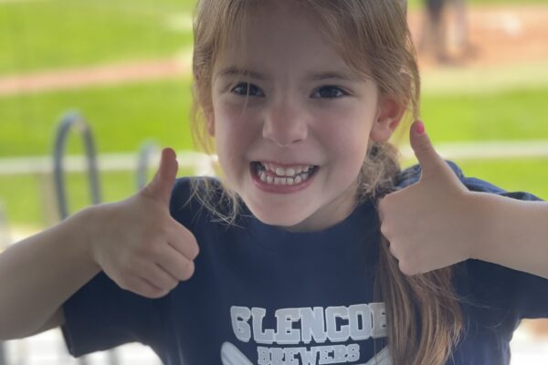 A young girl smiles and gives two thumbs up while wearing a navy blue Glencoe Braves Baseball shirt. She stands in front of a baseball field with players and green grass in the background.