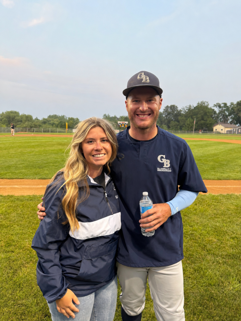 A woman and a baseball player, both smiling, stand together on a grassy baseball field. The player holds a water bottle, and the field and trees are visible in the background.