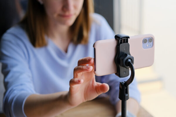 A person adjusts a smartphone on a small tripod, preparing to record or take a photo at a table. The background is softly blurred.