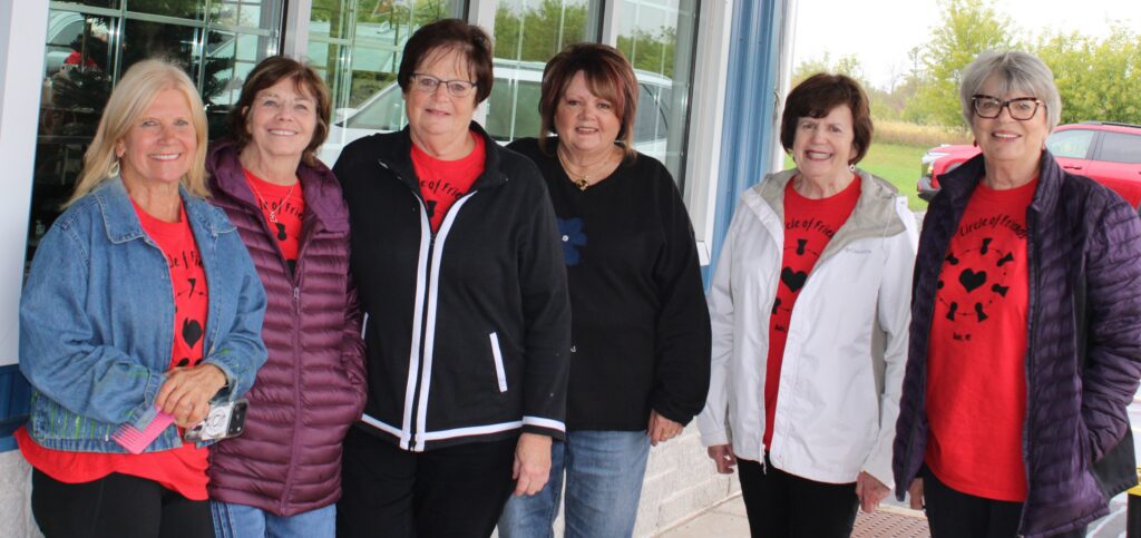 Six smiling women stand outside a building, wearing jackets and red shirts with a heart design. There are trees and parked cars in the background.