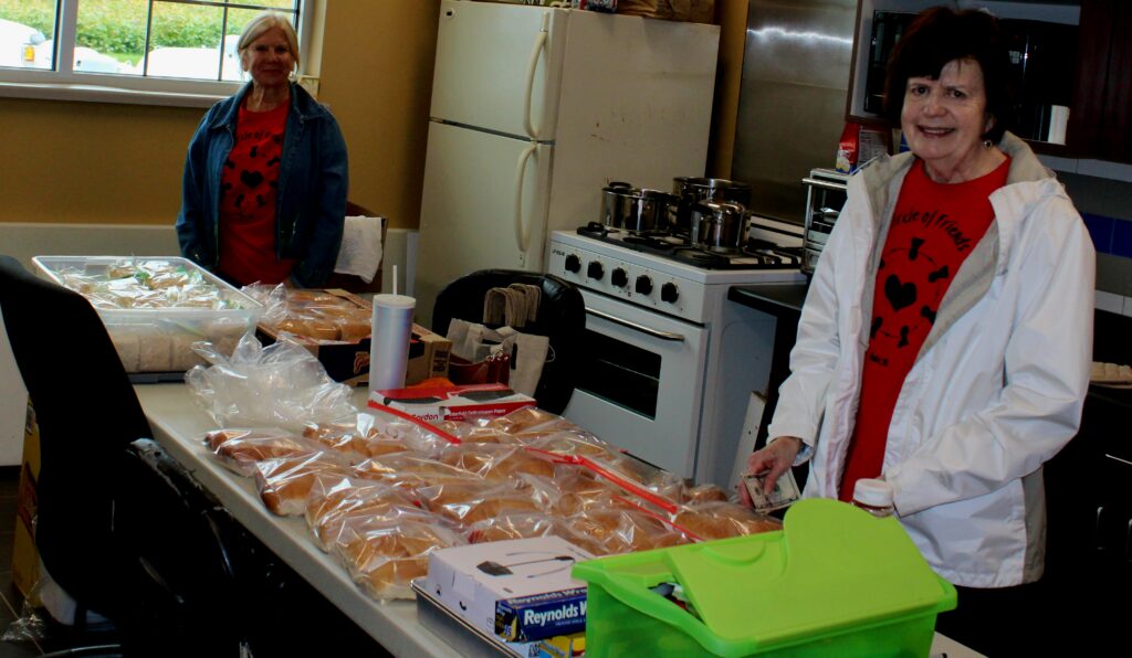 Two women in red shirts stand by a table filled with sandwich buns and supplies in a kitchen, preparing food. Pots are on the stove in the background.