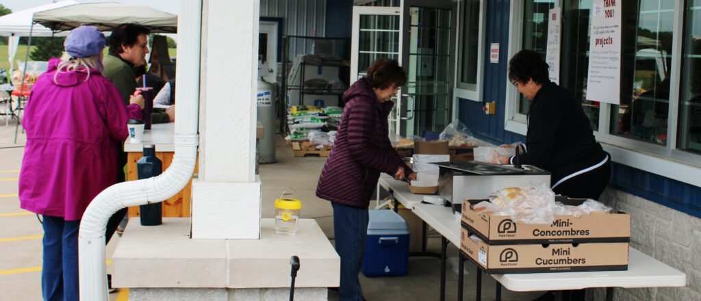 People stand outside by tables with food items. Two women are serving food from trays near boxes labeled Mini Cucumbers. Others wait nearby, some wearing jackets on a cloudy day. The setting appears to be a market or outdoor event.
