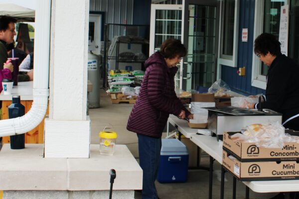 People stand outside by tables with food items. Two women are serving food from trays near boxes labeled Mini Cucumbers. Others wait nearby, some wearing jackets on a cloudy day. The setting appears to be a market or outdoor event.