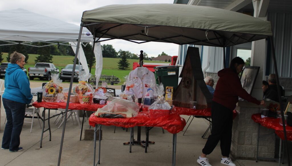 Outdoor market scene with tables covered in red plastic, displaying baskets and gifts under canopies. Two women and one man are present, one looking at items and another standing nearby.