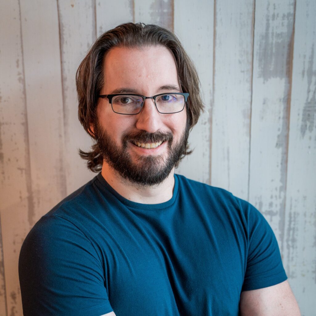 A smiling man with a beard and glasses wears a blue t-shirt as part of the marketing team.
