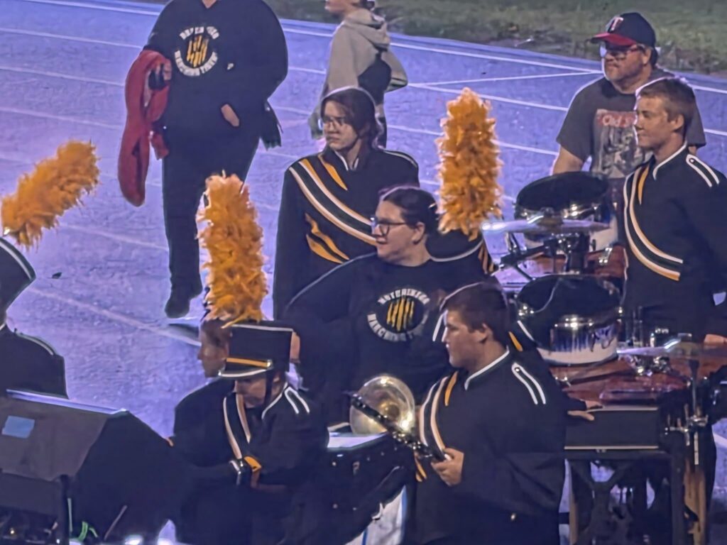 High school marching band percussionists in dark uniforms with yellow accents playing drums on a field.