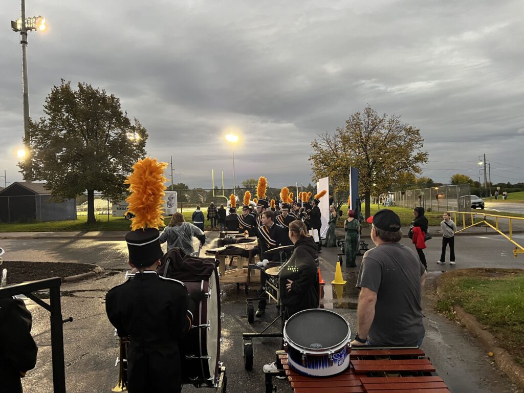 A high school marching band in black uniforms with yellow plumes practices on an asphalt field under stadium lights.