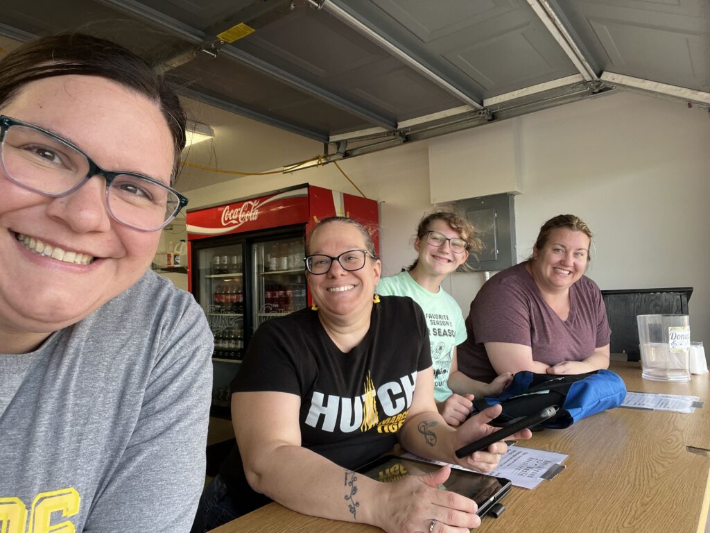 Four women smiling and posing for a selfie at a wooden table inside a garage.