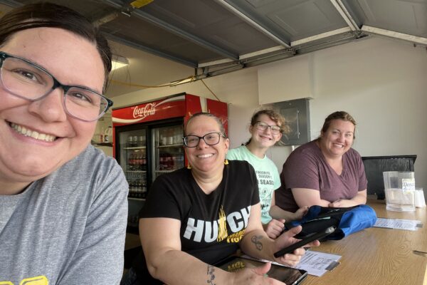 Four women smiling and posing for a selfie at a wooden table inside a garage.