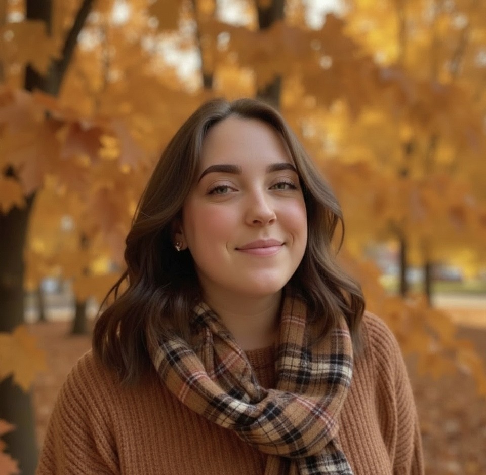 A young marketing team member wearing a brown sweater and plaid scarf poses in front of autumn trees with golden leaves.